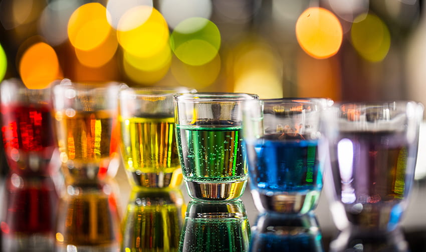 A lineup of rainbow-colored liquids in shot glasses at a bar