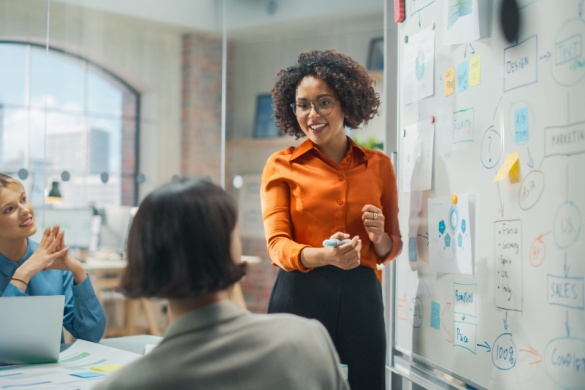 A woman stands in front of a whiteboard sharing her presentation with colleagues