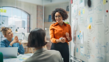 A woman stands in front of a whiteboard and presents her project plans to fellow employees